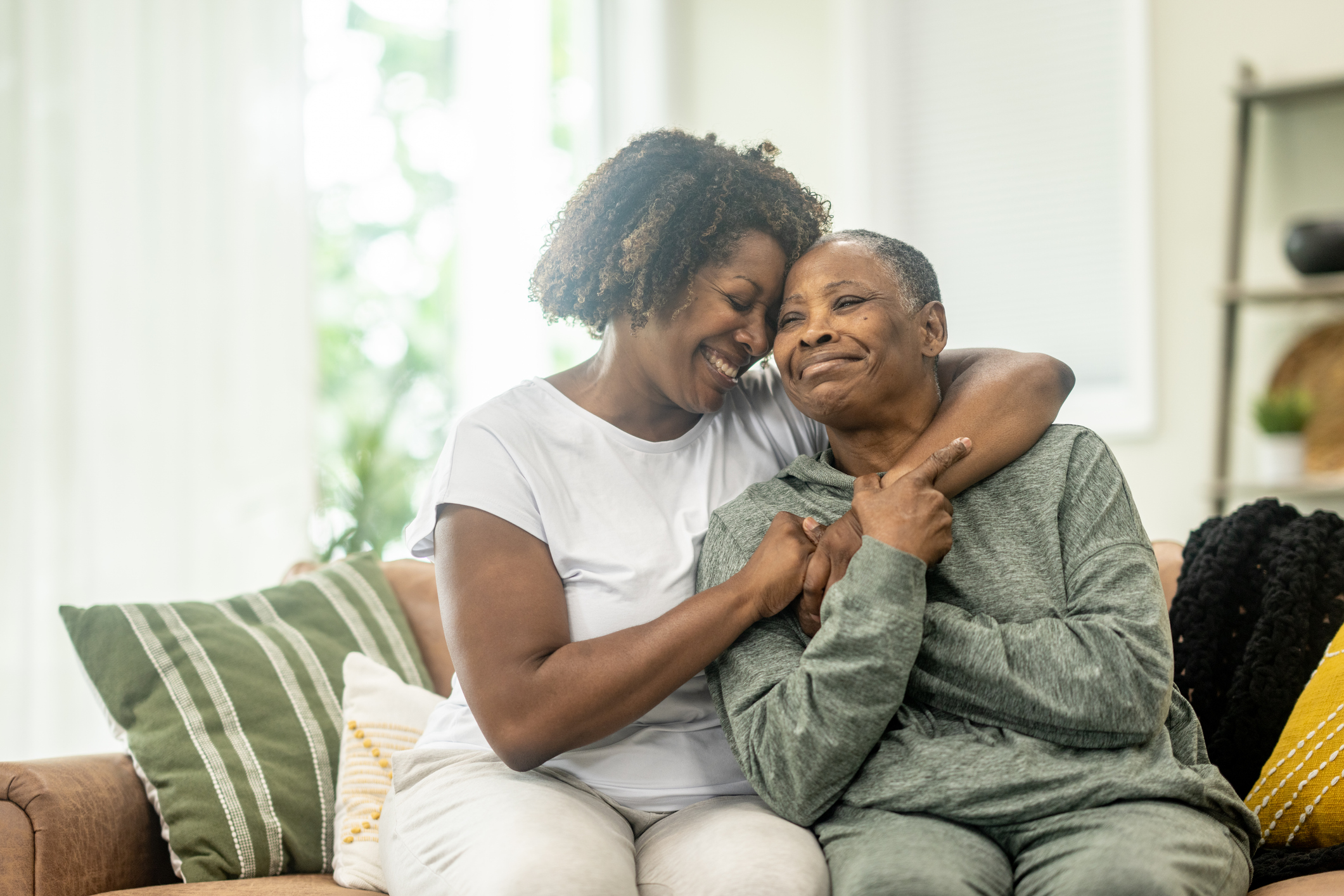 A middle-aged woman of African descent wraps her arm around her Mother as she embraces her and they pose for a portrait. They are both dressed comfortably and are laughing as they enjoy their time together.