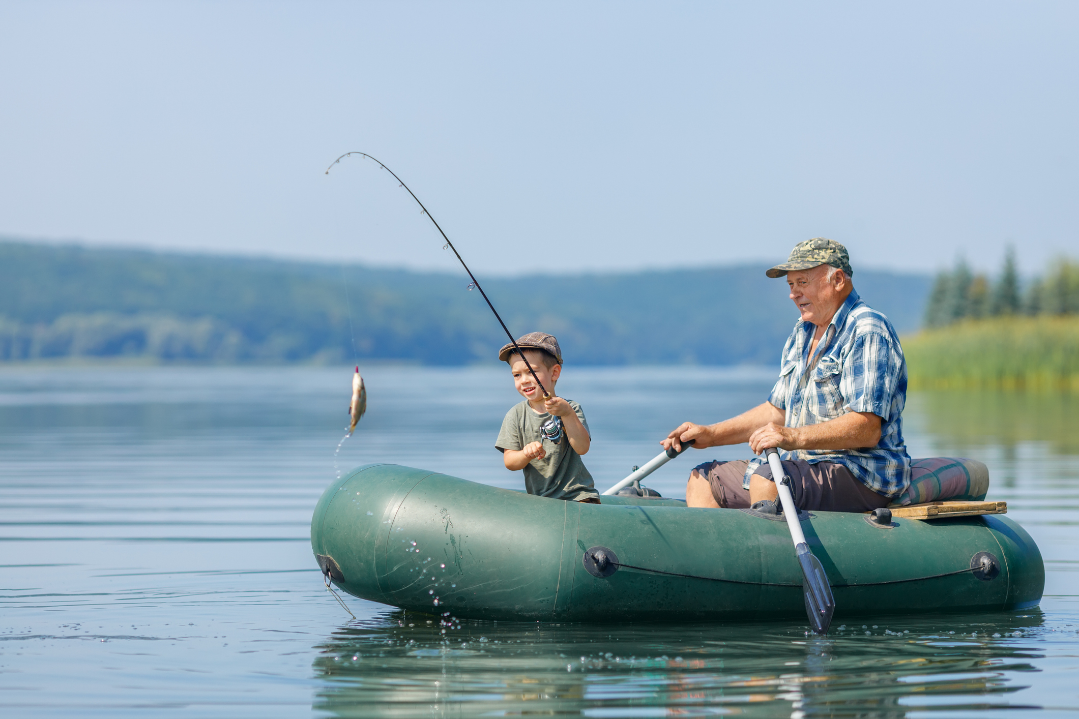 A calm large lake with a green dinghy type boat on the water. A young boy is in the boat with a fishing rod and a fish attached to the end whilst an older man rows the boat.